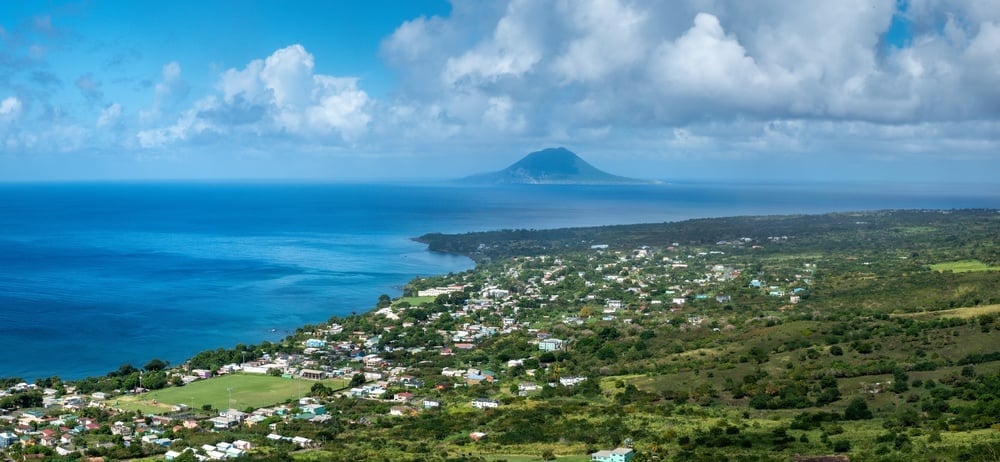 Aerial view of a tropical island with blue ocean, volcanic mountain in distance, green vegetation, and scattered coastal settlements