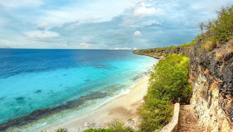Tropical beach with turquoise waters, white sand, and limestone cliffs covered in green vegetation under a blue sky with white clouds
