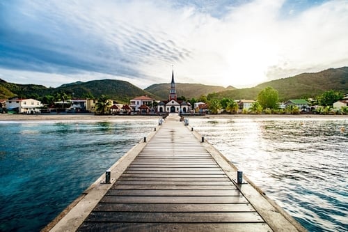Wooden pier extending into turquoise water toward a waterfront town with a church spire and mountains under blue sky