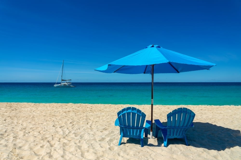 Two blue Adirondack chairs under a blue umbrella on a sandy beach with turquoise water and a sailboat in the background