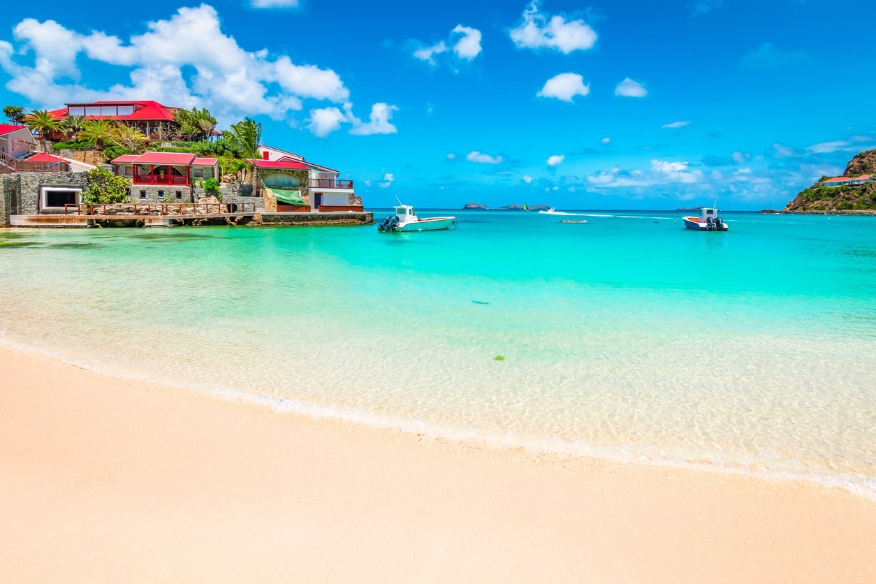 Tropical beach resort with turquoise water, white sand, red-roofed villas, and boats under clear blue sky