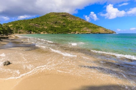 Tropical beach with turquoise water, sandy shore, and lush green hillside under blue sky with clouds