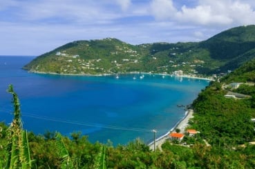 Aerial view of a lush, forested coastline with turquoise bay waters, white sailboats, and hillside homes nestled among green vegetation
