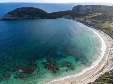 Aerial view of a crescent beach with turquoise waters, rocky outcrops, and forested headlands along a rugged coastline