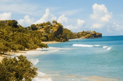 Tropical beach with turquoise water, white sand, lush green vegetation, and rocky headland under blue sky with white clouds