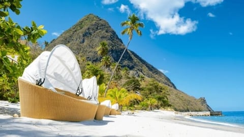Luxury beach pod on sandy shore with white sailboat canopy, volcanic mountain backdrop, and turquoise ocean under blue sky