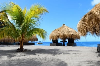 Tropical beach scene with palm trees, thatched-roof huts, blue ocean, and white sand under clear sky