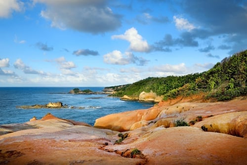 Scenic coastal landscape with sandy beach, smooth boulders, blue ocean, forested cliffs, and partly cloudy sky