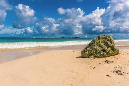 Moss-covered rock on sandy beach with turquoise ocean, white waves, and blue sky with clouds in background