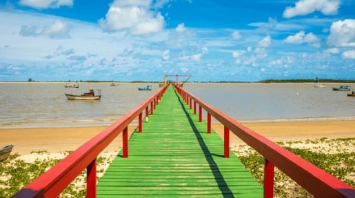 Green wooden boardwalk with red railings extending into calm turquoise water under blue sky with white clouds