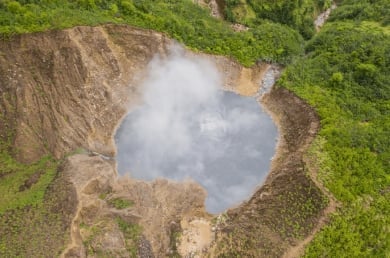 Aerial view of a circular crater lake surrounded by tan rocky walls and dense green forest vegetation