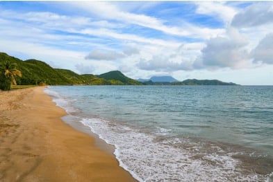 Sandy beach with turquoise waves, forested hills, and blue sky with clouds on the horizon