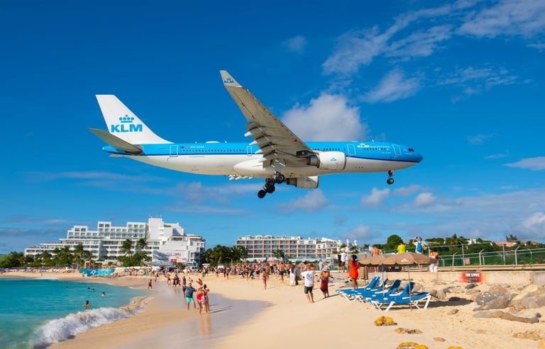 KLM airplane flying low over a crowded beach with turquoise water and coastal buildings