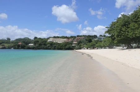 Tropical beach with turquoise water, white sand, and lush green hillside with buildings under blue sky