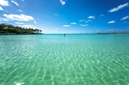 Clear turquoise tropical waters with rippling patterns, green forested coastline, and a lone person in the distance under blue sky with white clouds