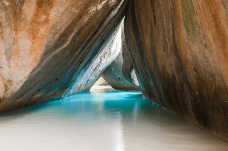 Turquoise water flowing between massive boulders, with a small white sandy beach visible through the rocky archway