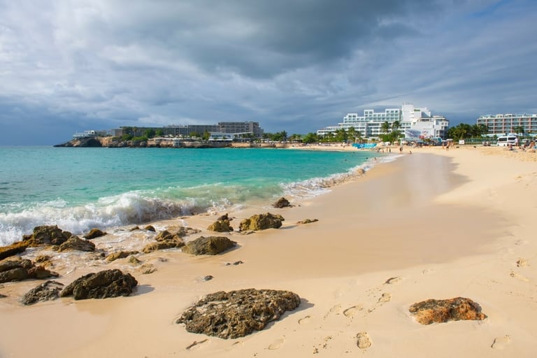 Scenic coastal beach with turquoise waters, sandy shores, dark rocks, and white resort buildings under cloudy skies