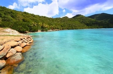 Turquoise tropical beach with rocky shoreline, lush green hills, and blue sky with white clouds