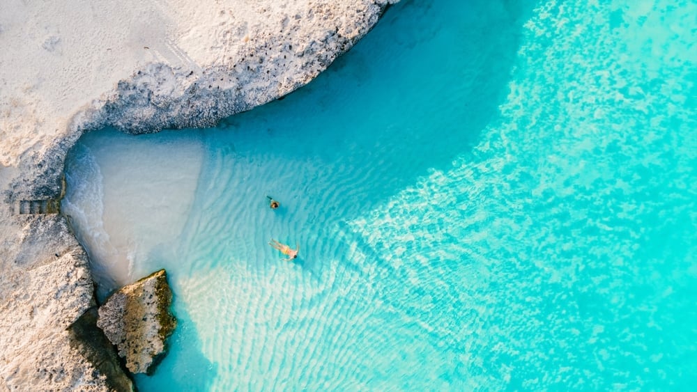 Aerial view of turquoise beach water with sandy shore and small swimmers in crystal clear shallow bay