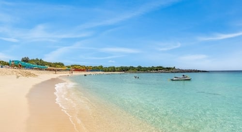 Scenic sandy beach with turquoise water, white-sailed boat, and beachgoers under clear blue sky
