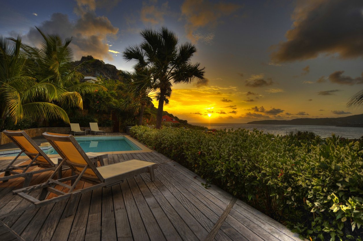 Two lounge chairs on a wooden deck overlooking a tropical beach at sunset with palm trees and ocean view