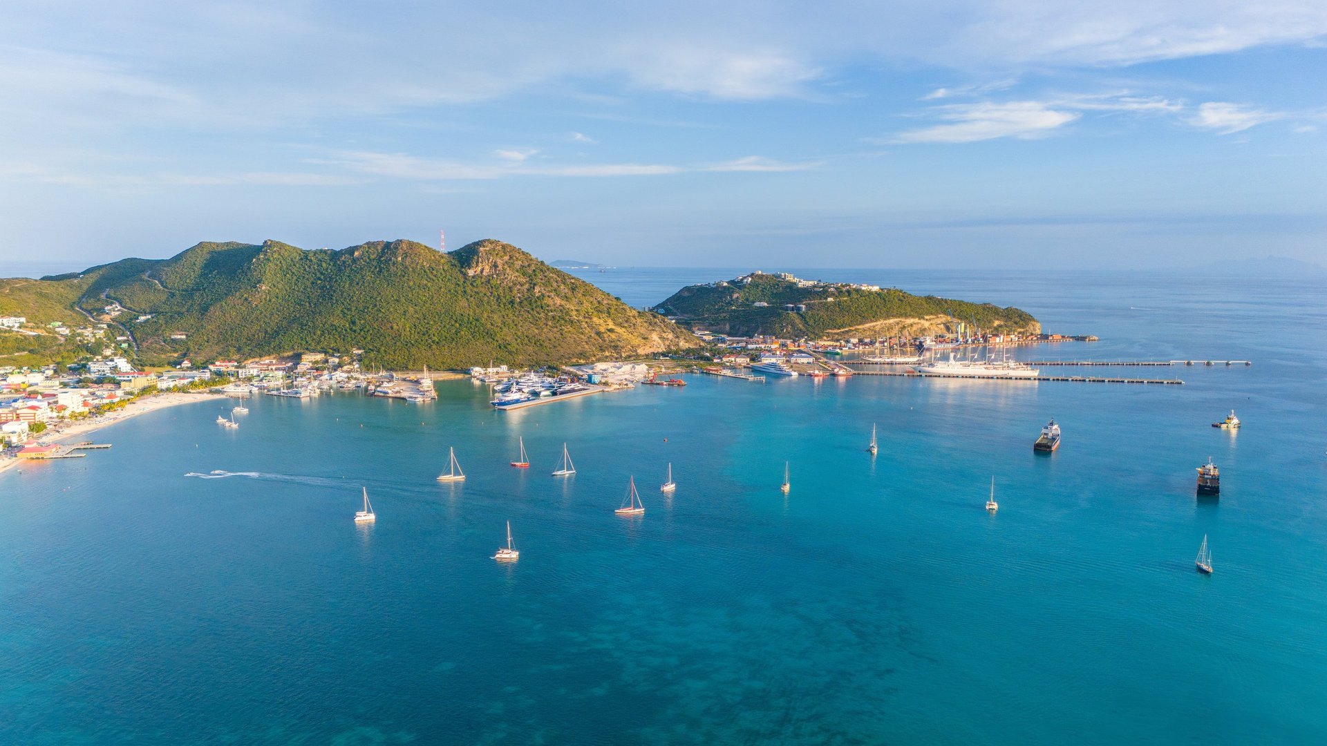 Aerial view of a Mediterranean coastal town with turquoise waters and numerous sailboats anchored in the bay, surrounded by forested hills