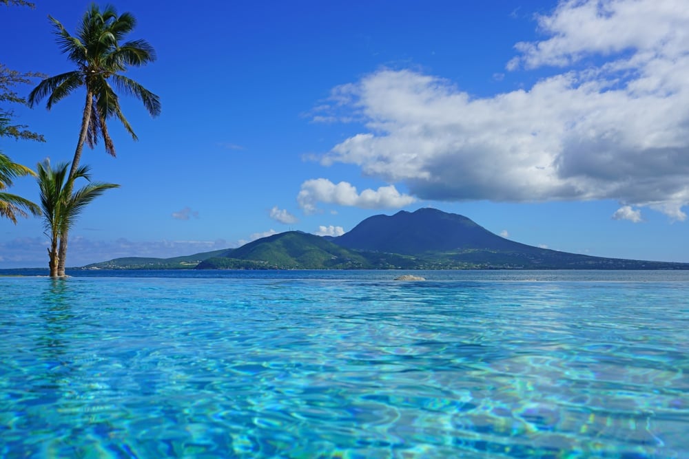 Palm tree in turquoise water with volcanic island mountain in the distance under blue sky